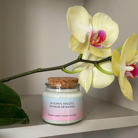 Candle with a pink label next to a yellow orchid on a white surface